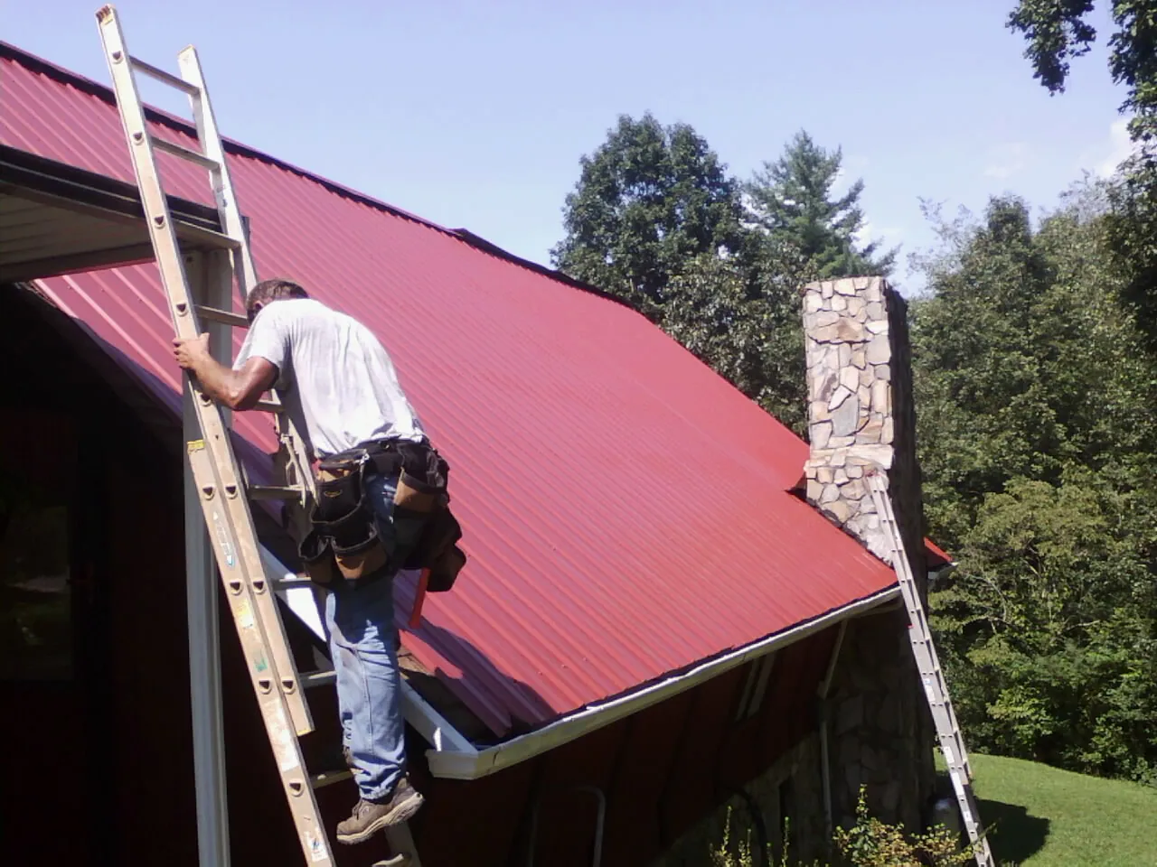 Roofer on a ladder beside a red metal roof and stone chimney — Soffit & Fascia Repair by Roofing Brothers in Emington